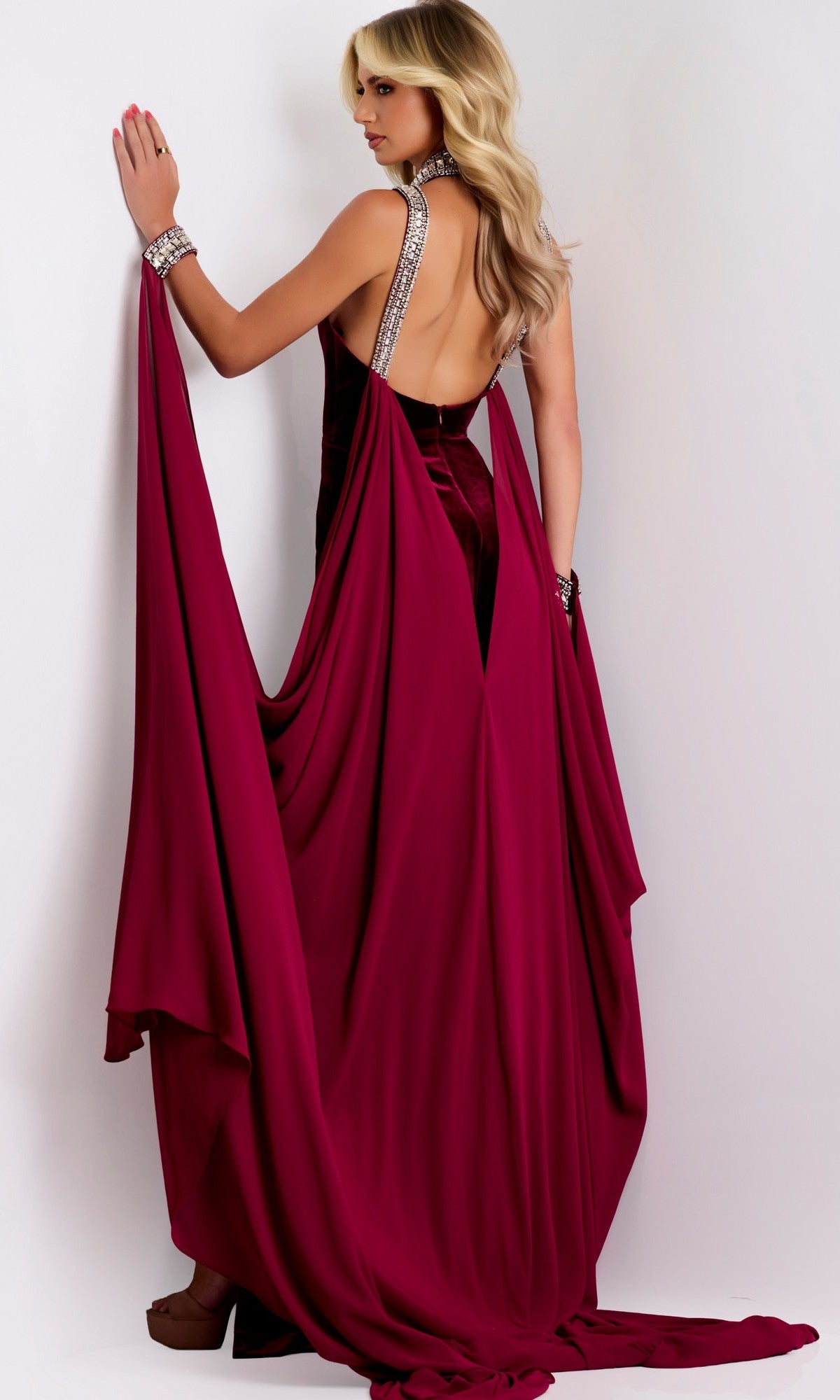 A woman wearing the Jovani 43699 long prom dress—featuring a backless velvet halter design with draped fabric and a high-leg slit—stands against a white background, facing away from the camera.